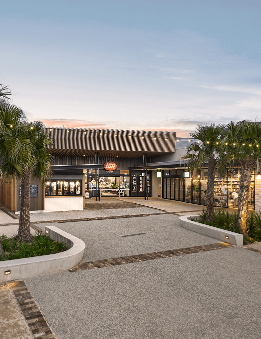 Modern IGA supermarket entrance at dusk with palm trees and string lights.