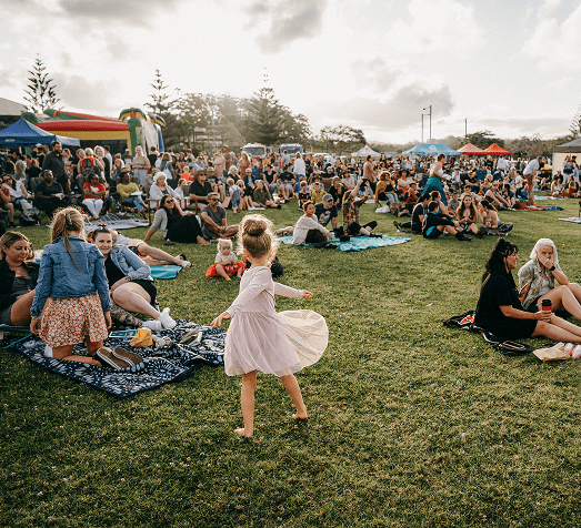 Crowd of people seated on grass at an outdoor event with children playing and tents in the background during daytime.