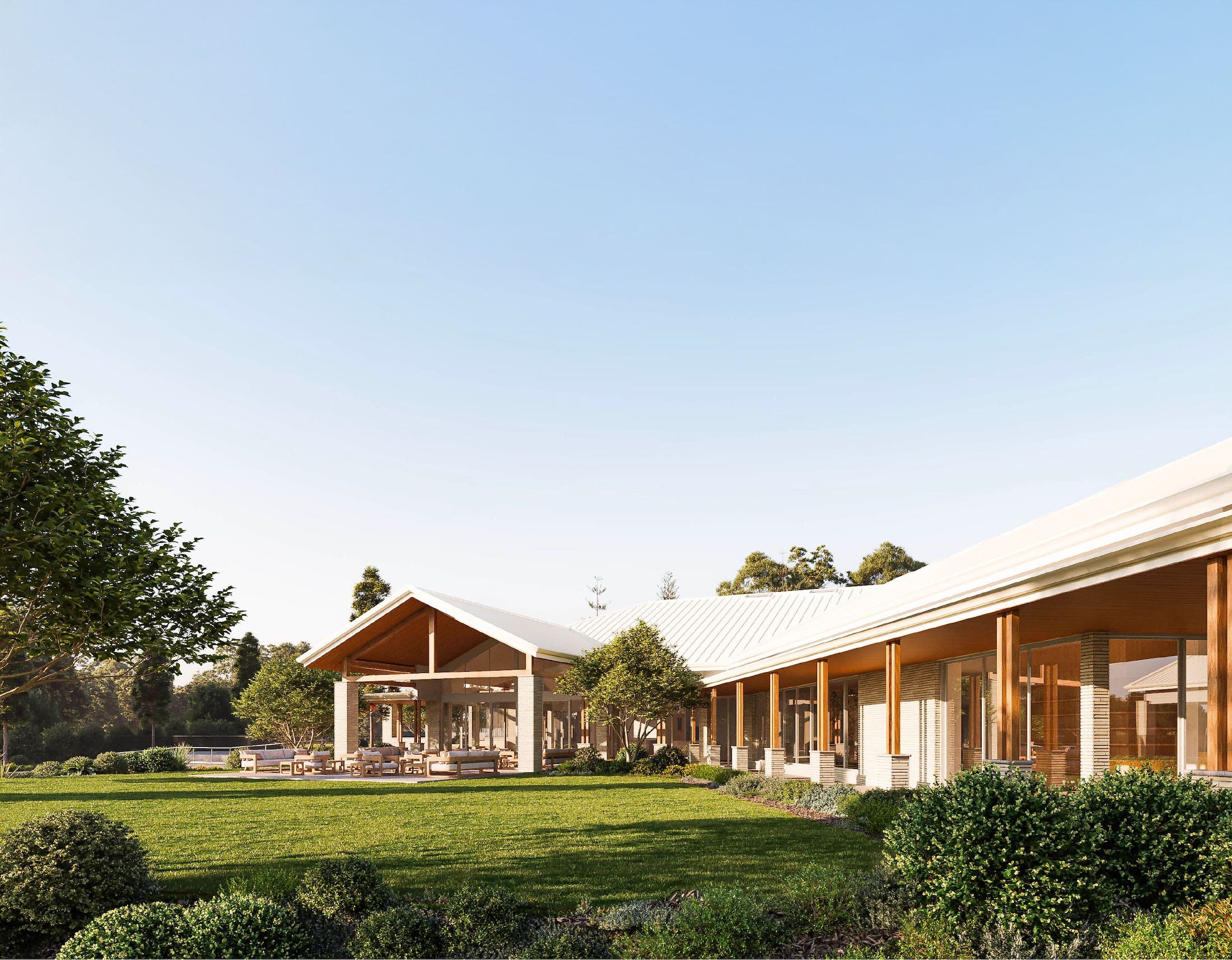 Modern single-story building with large glass windows, wooden columns, and outdoor seating area surrounded by green lawn and bushes under a clear blue sky.