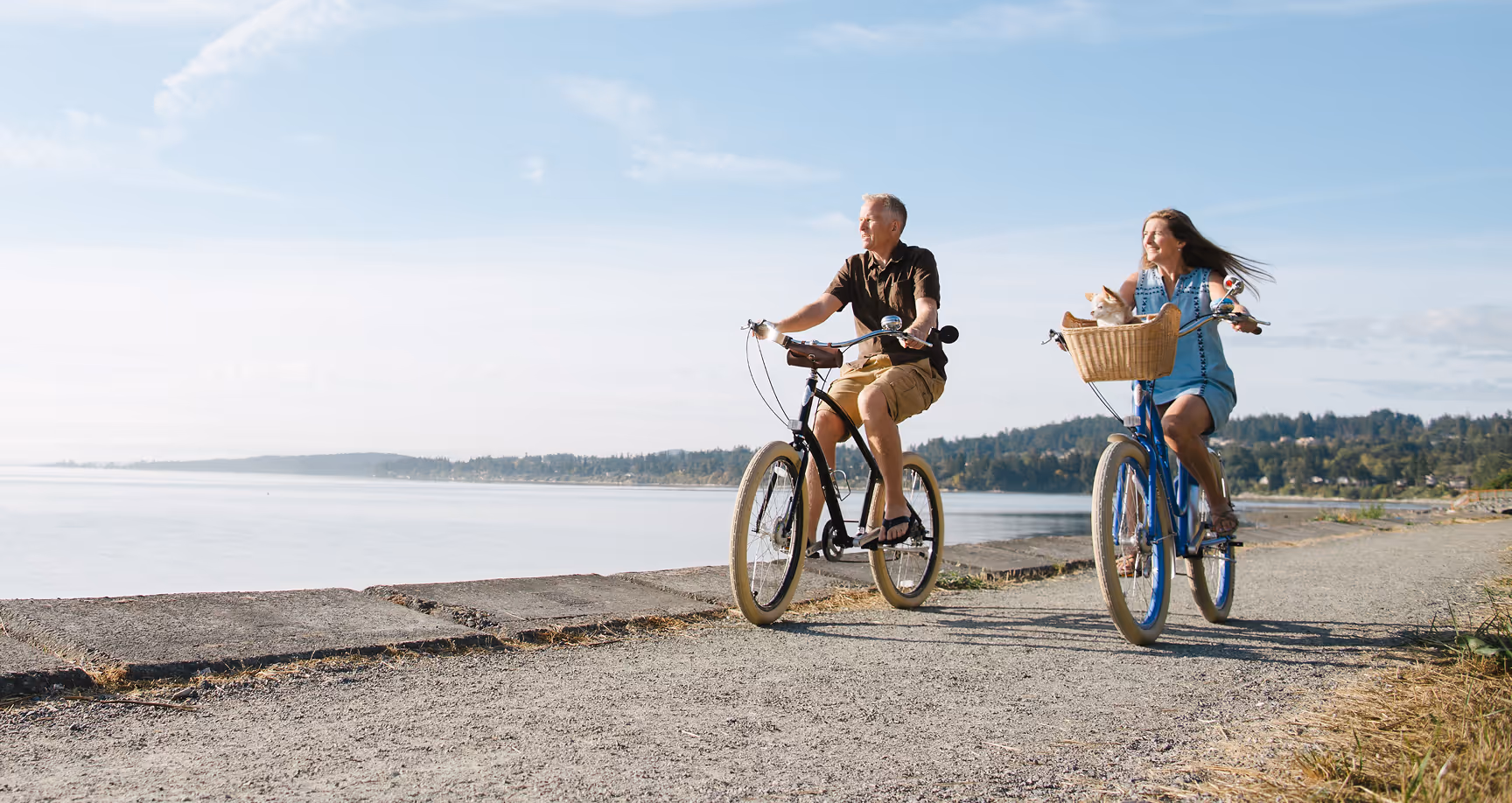 Man and woman riding bicycles on a seaside path, with a cat in the woman's bike basket.