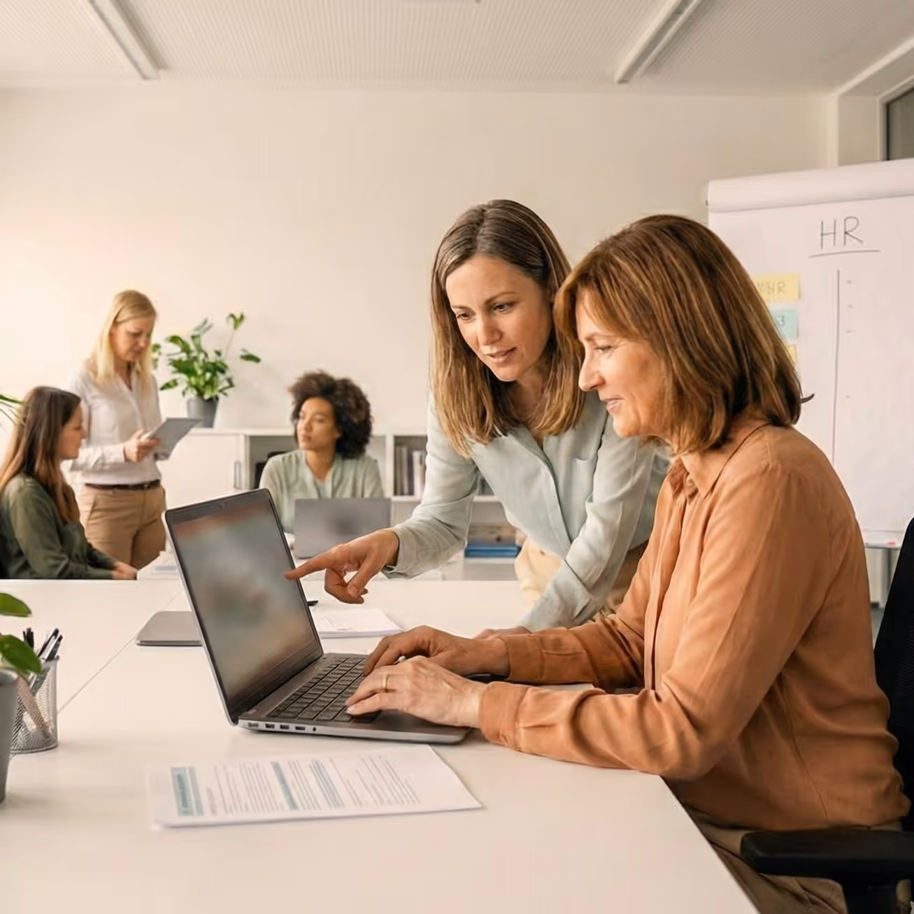 Two women collaborating over a laptop in a bright office with others working in the background near an HR flip chart.