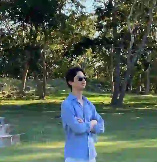 Person wearing a blue shirt and sunglasses standing outdoors on grass with arms crossed, surrounded by trees.