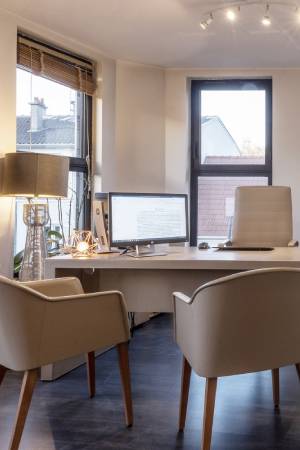 Bureau moderne avec deux fauteuils beige, un ordinateur, une lampe et deux grandes fenêtres avec vue sur des toits.