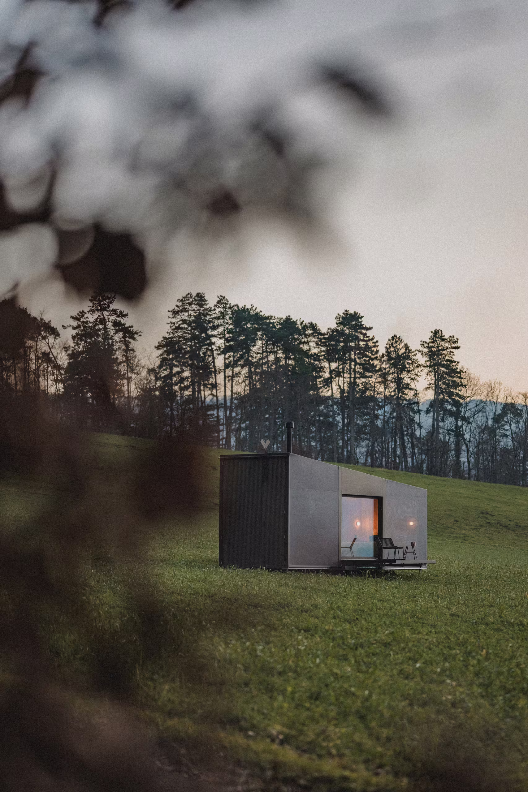 Tiny house di lusso SNUUG illuminata al crepuscolo in un prato in Svizzera, un concetto di hotel itinerante a 4 stelle senza impronta ecologica.