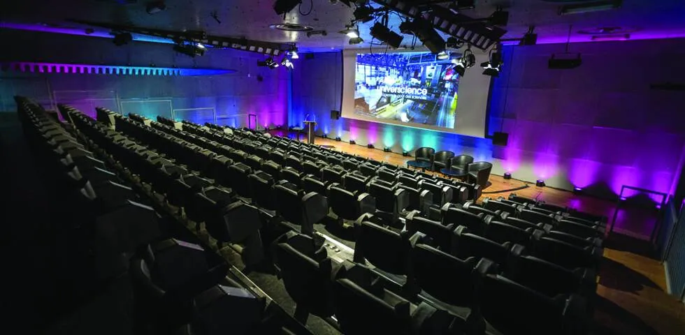 Empty auditorium with rows of black chairs facing a stage with four chairs and a podium, illuminated by purple and blue lights, with a large screen displaying a science presentation.