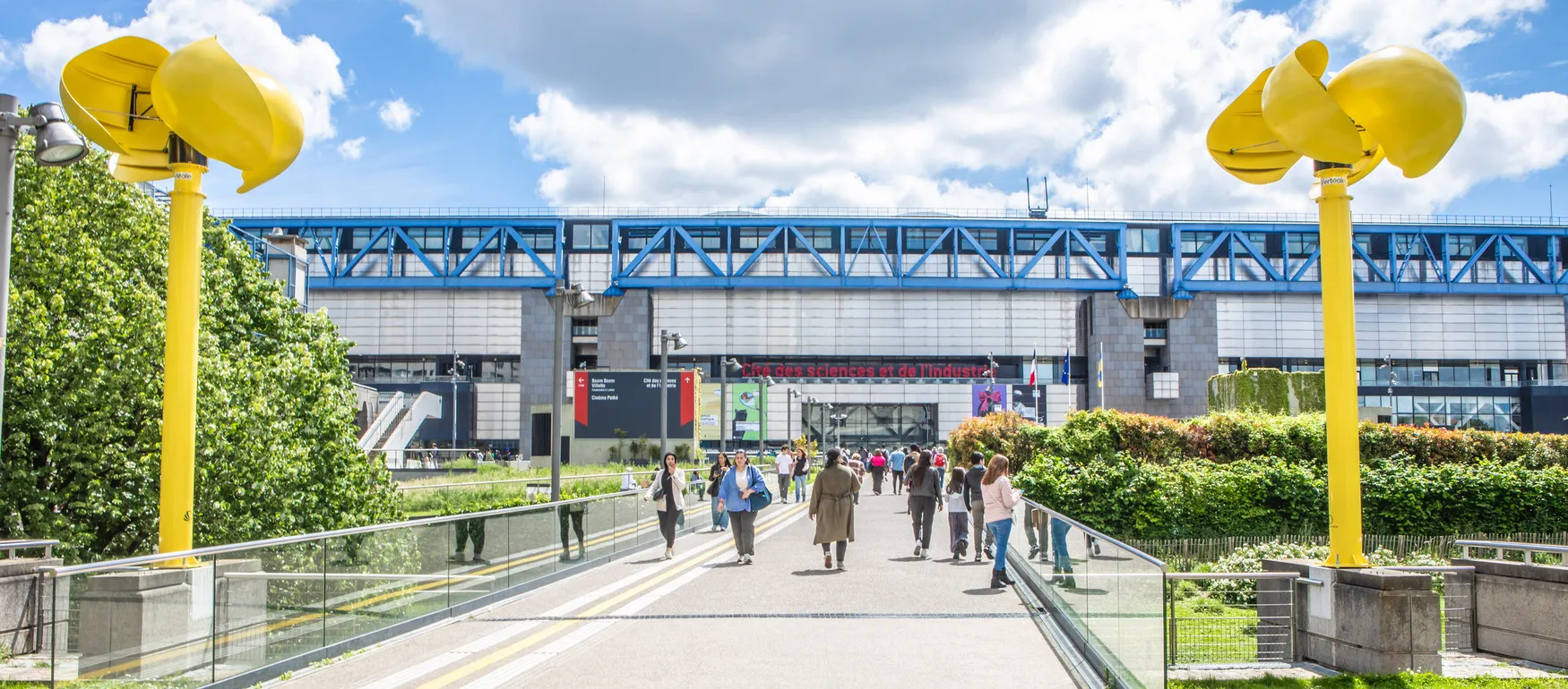 Walkway with people leading to a modern building labeled Cité des sciences et de l'industrie with yellow wind turbines on both sides and green bushes.