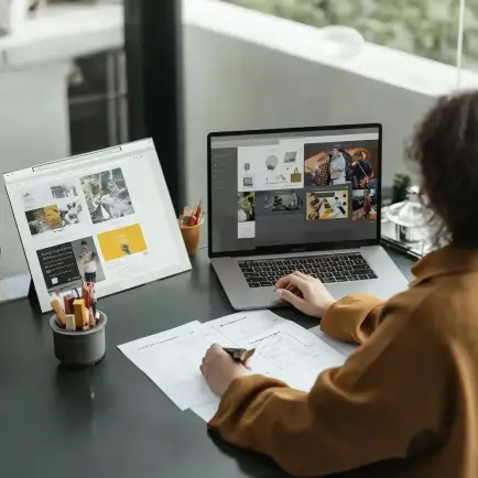 Person sketching website designs on paper at a desk with a laptop and monitor displaying visual content.