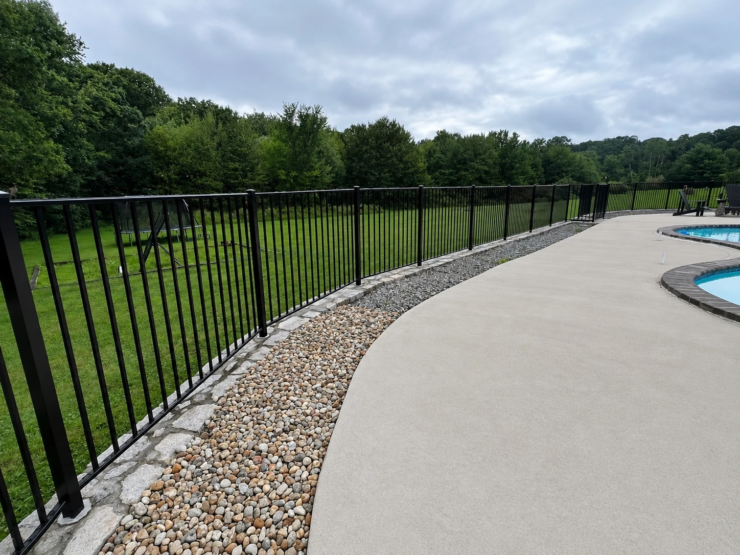 Black aluminum pool fence installed beside a curved concrete patio with stone border landscaping.