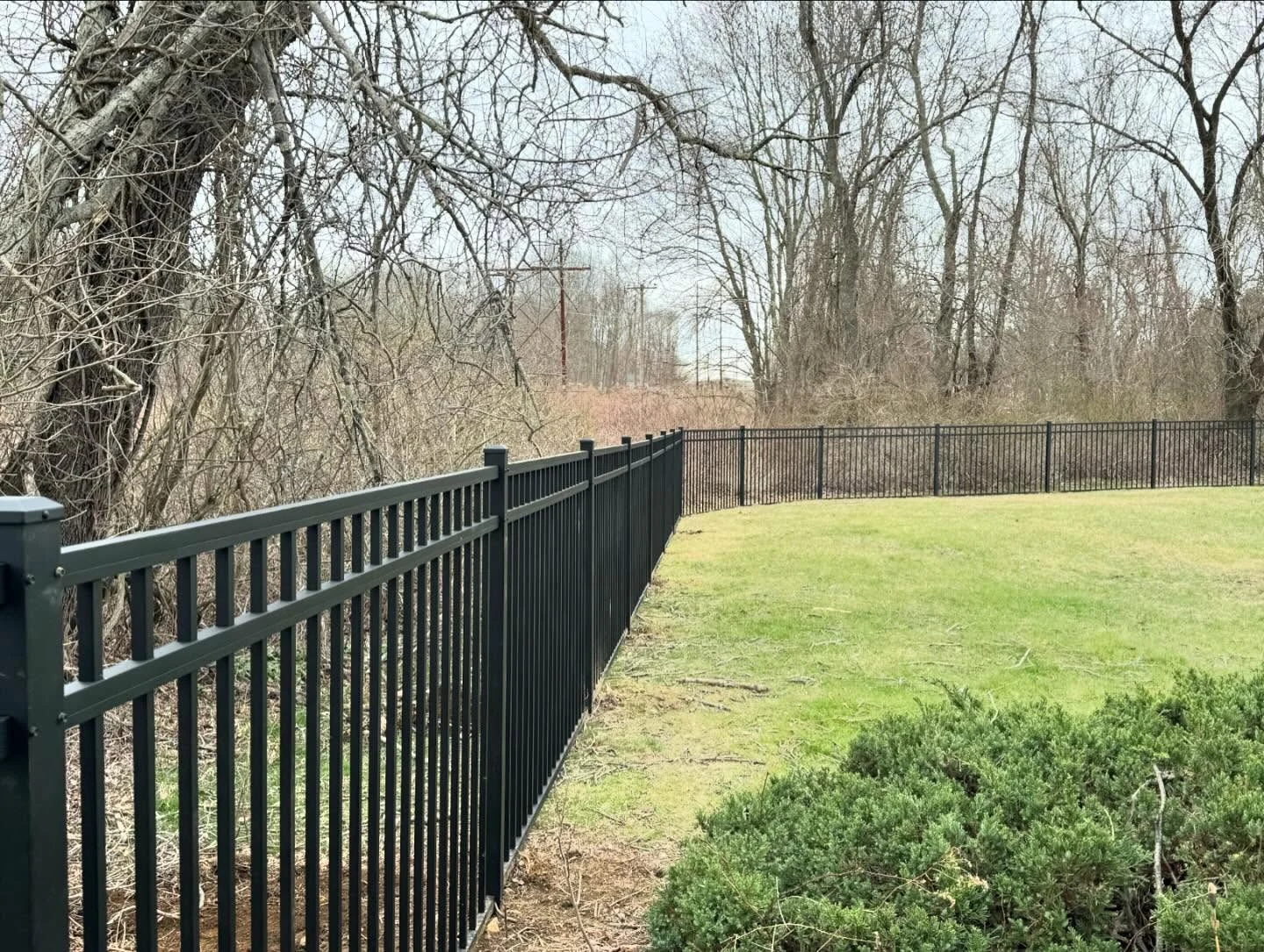 Three-rail aluminum perimeter fence installed along a wooded residential property line.