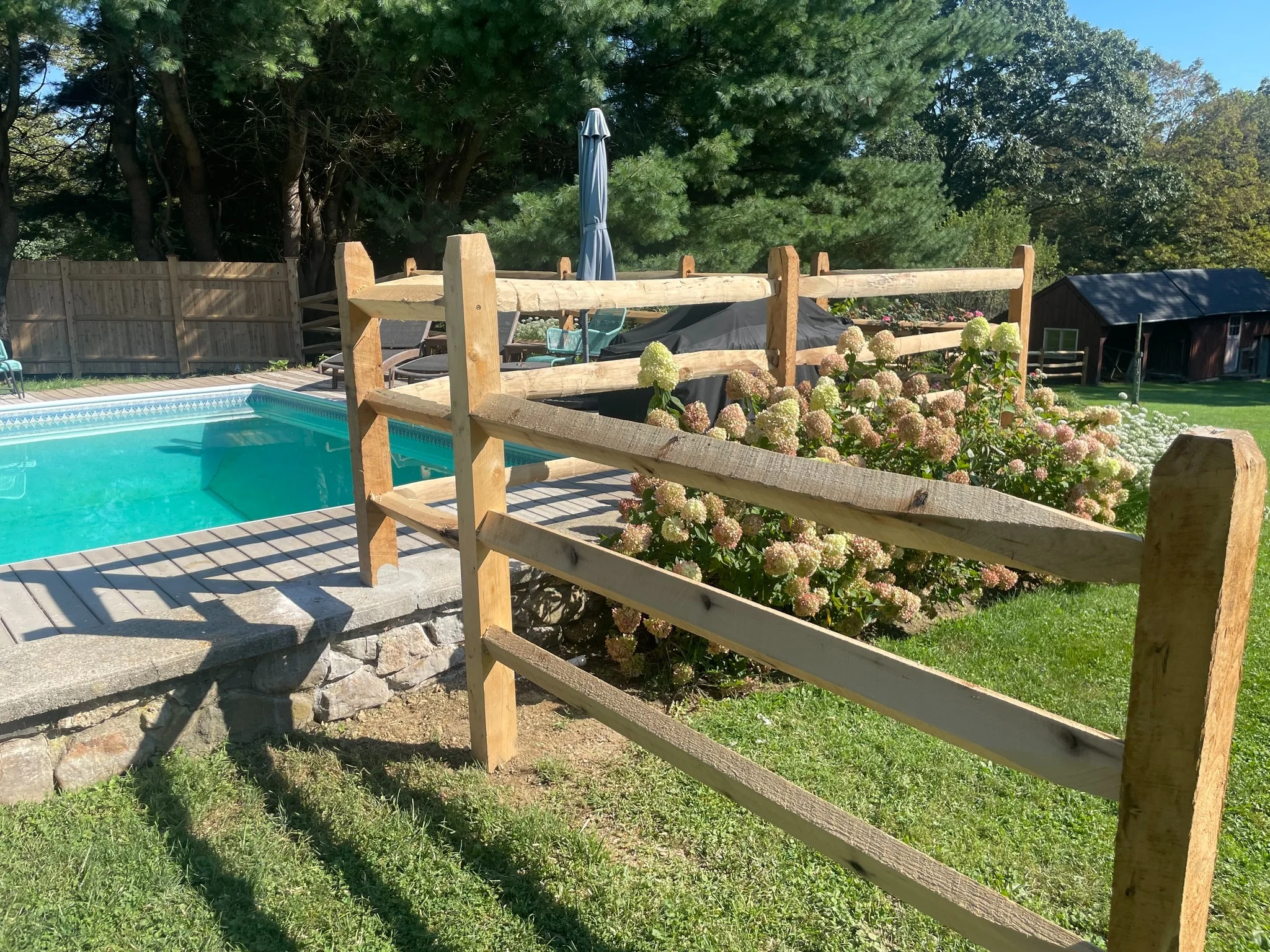 Natural wood three-rail fence surrounding a backyard pool with hydrangeas and lawn.