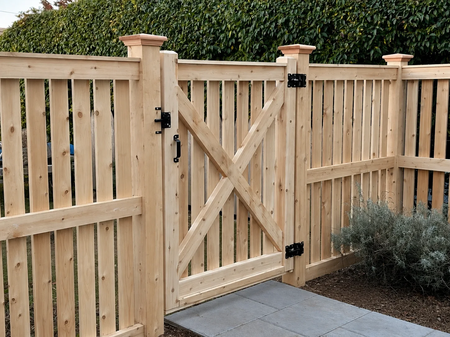 Natural wood picket gate with cross bracing and black hardware beside a paved walkway.