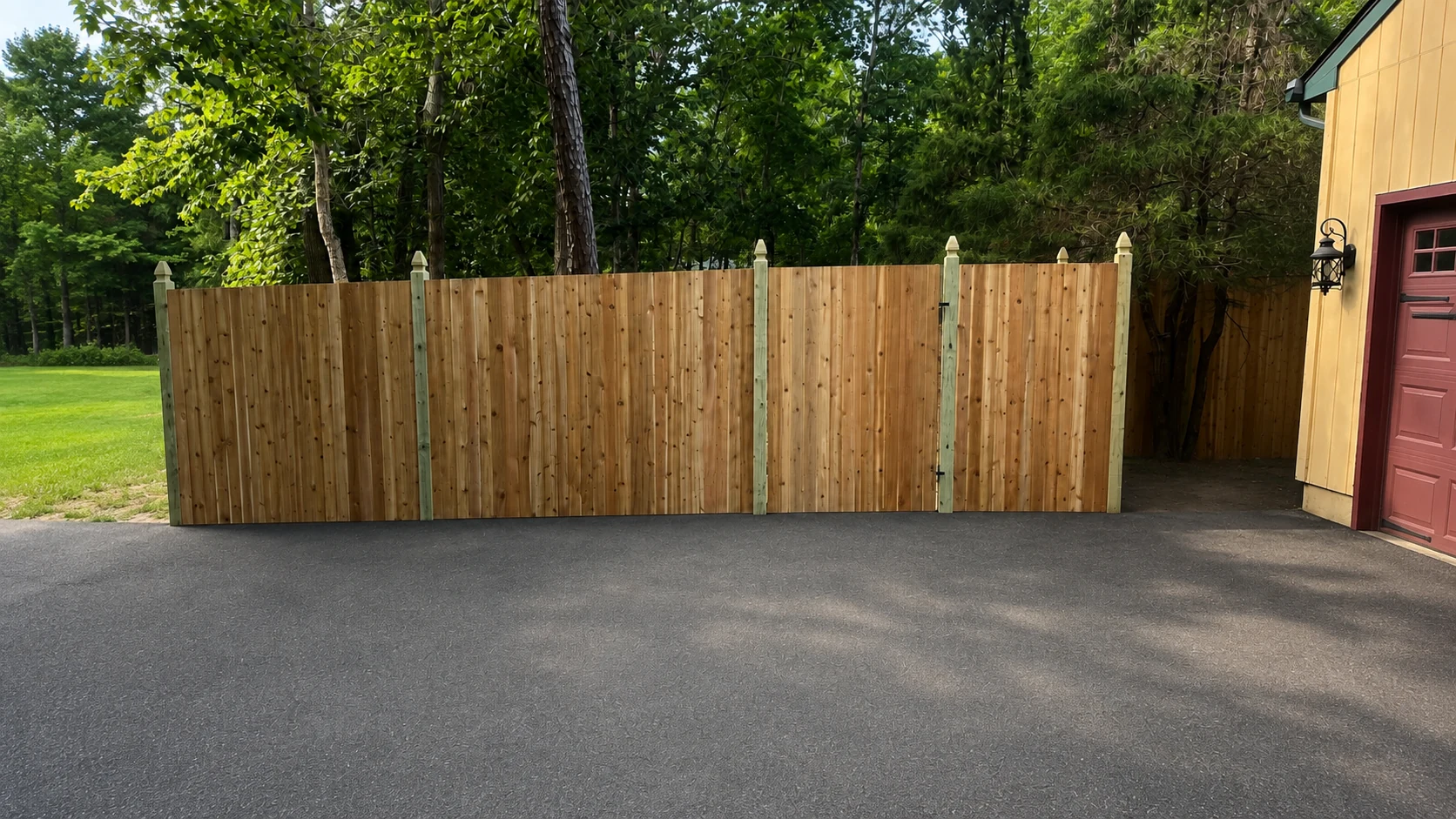 Tall wood privacy fence and gate installed beside a driveway with mature trees behind it.