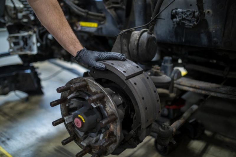 Mechanic wearing black gloves inspecting heavy-duty truck brake assembly, showing brake drum, pads, and wheel hub components inside a maintenance workshop.