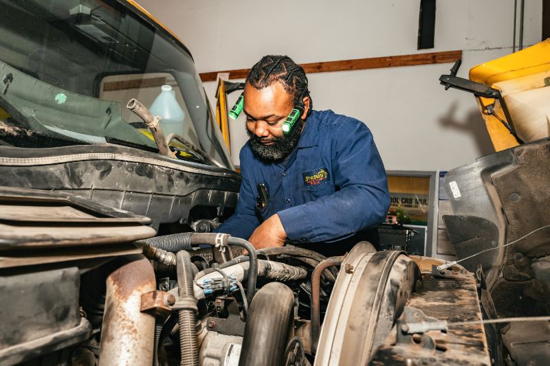 Diesel mechanic in blue uniform working on truck engine inside repair shop, focused on maintenance, wearing flashlight headlamps for better visibility.