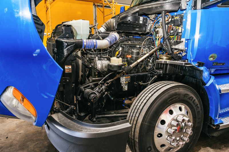 Open hood of a blue semi-truck showing detailed diesel engine components, hoses, and wiring inside a repair shop environment.