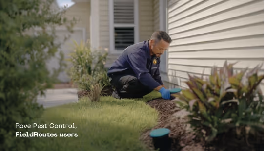 Man wearing gloves inspecting soil near a house foundation with plants around.