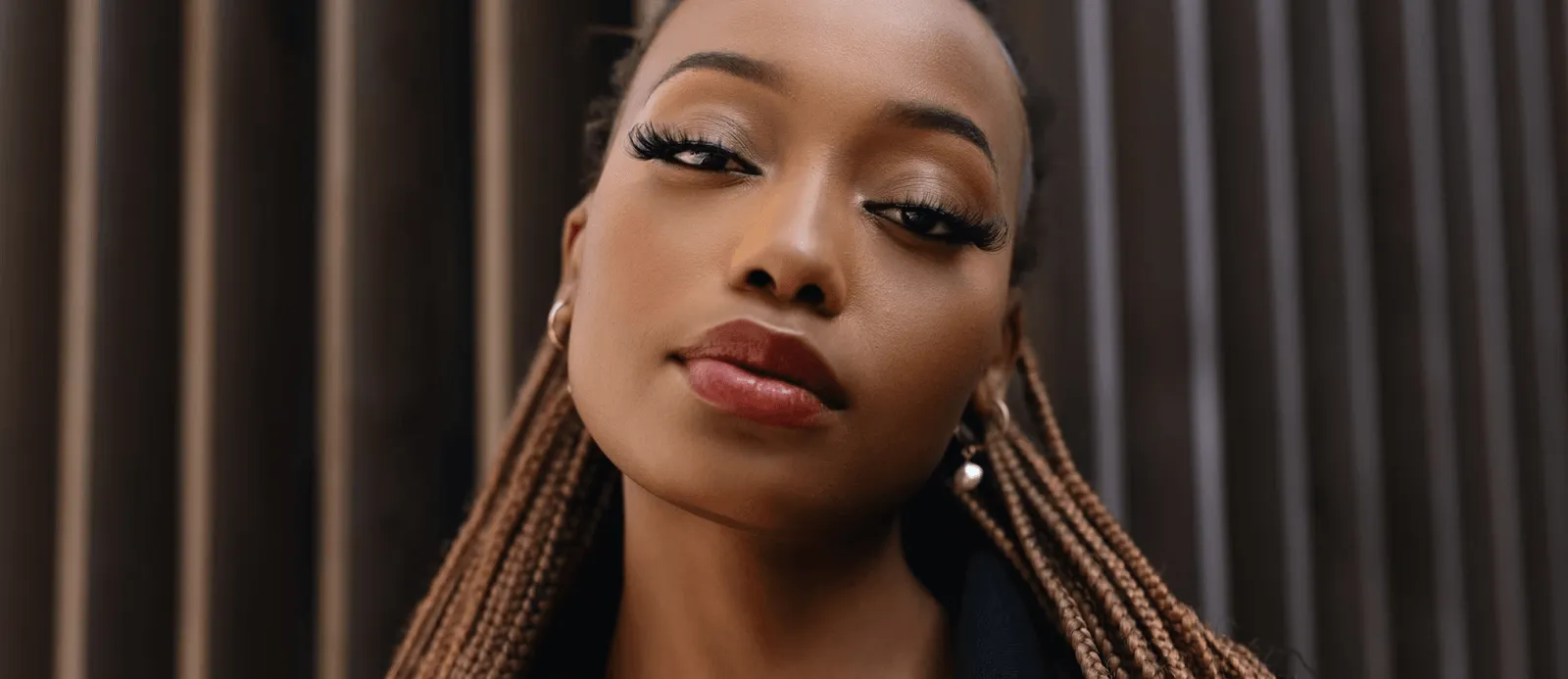 Close-up portrait of a woman with long braided hair, wearing pearl earrings and red lipstick, against a dark vertical background.