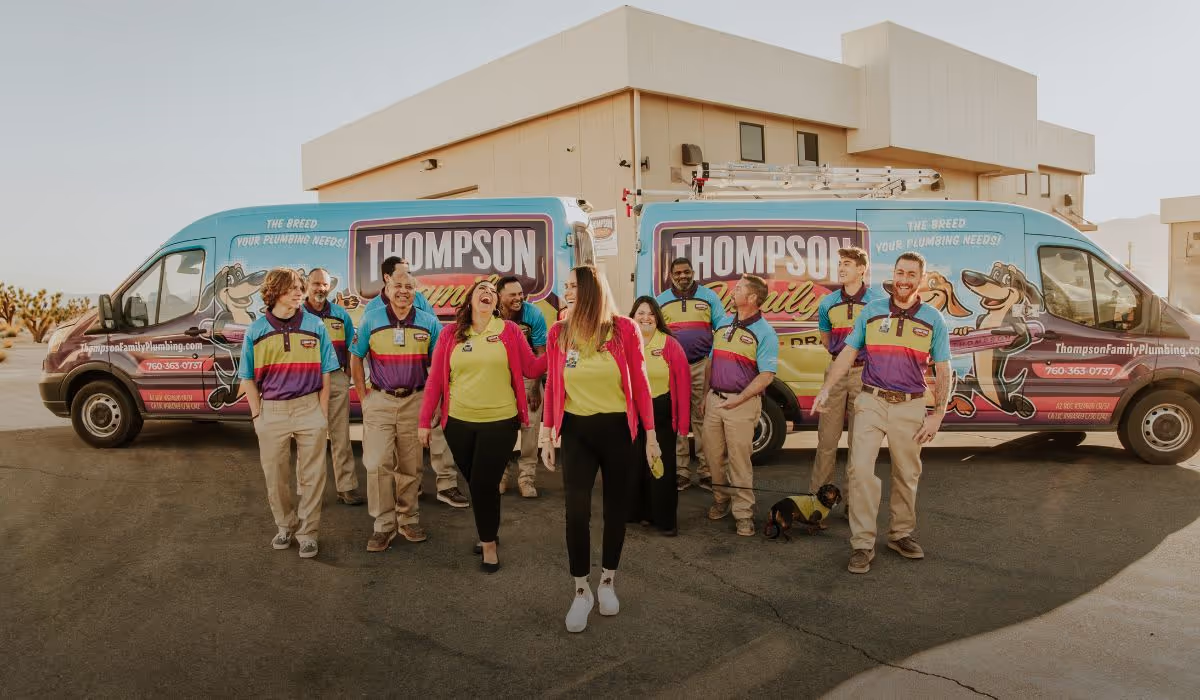 Group of Thompson Family Plumbing employees standing and walking in front of two company vans with colorful branding.