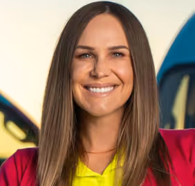 Smiling woman with straight brown hair wearing a red blazer and yellow shirt, standing outdoors.