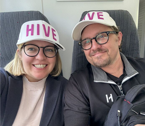 A smiling man and woman wearing white caps with the word 'HIVE' seated on patterned chairs.