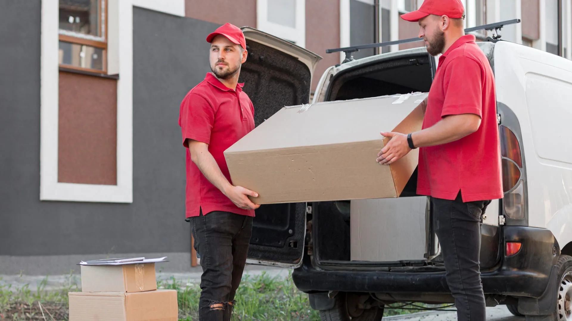 Two movers in red shirts and caps carrying a large cardboard box by a white van.