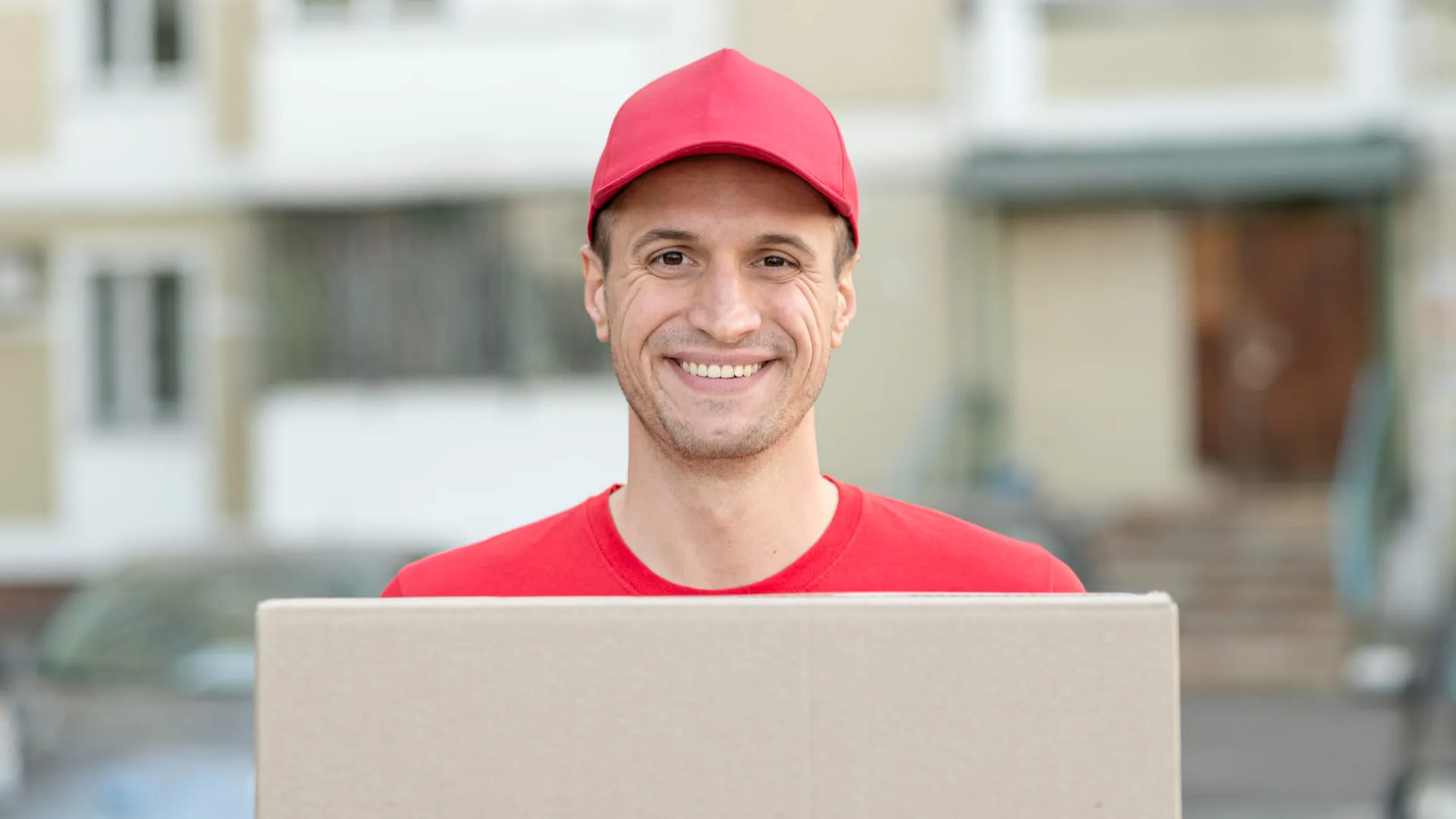 Smiling delivery man in red cap and shirt holding a cardboard box outdoors.