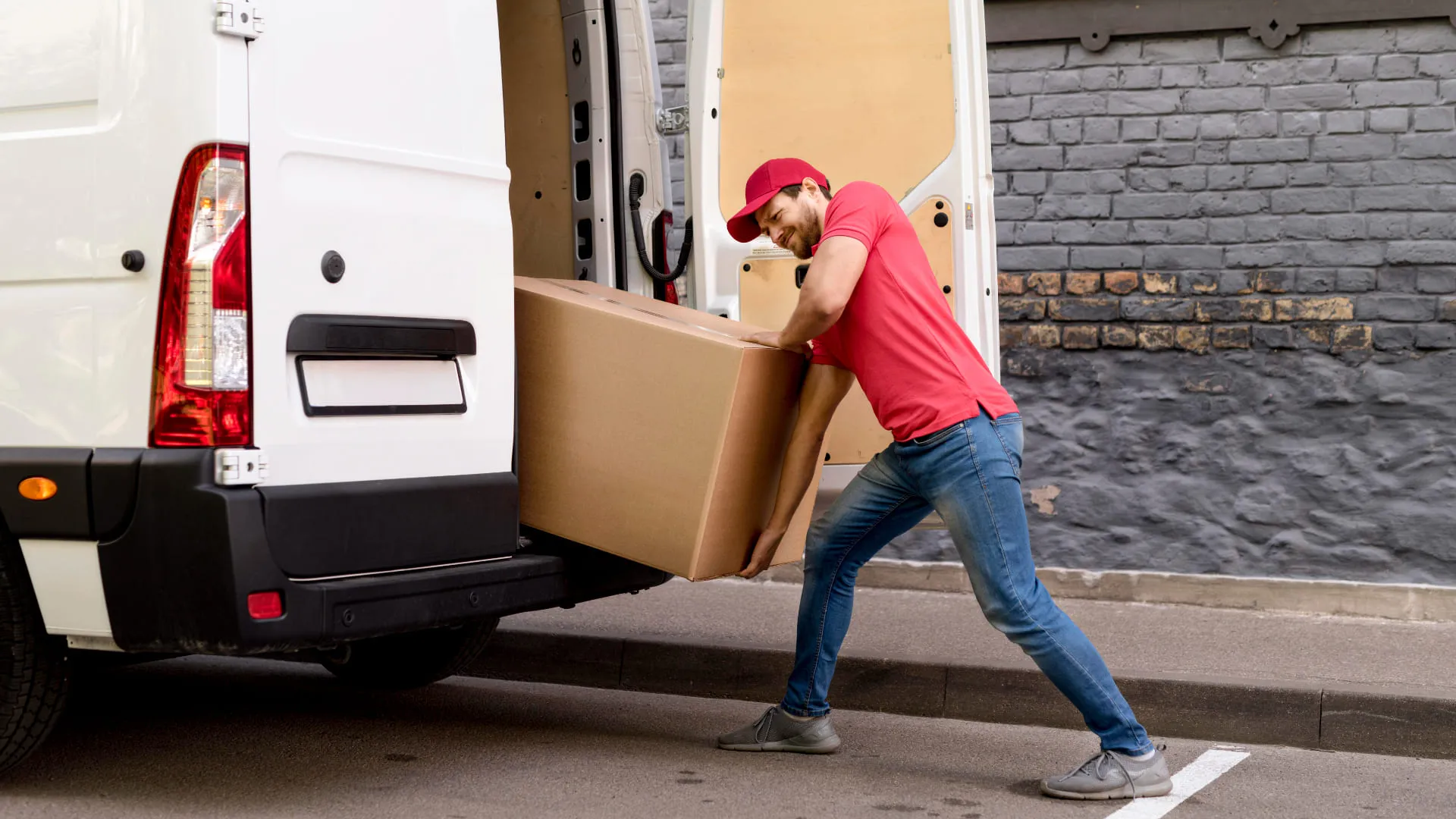 Man in red shirt and cap loading a large cardboard box into a white delivery van.