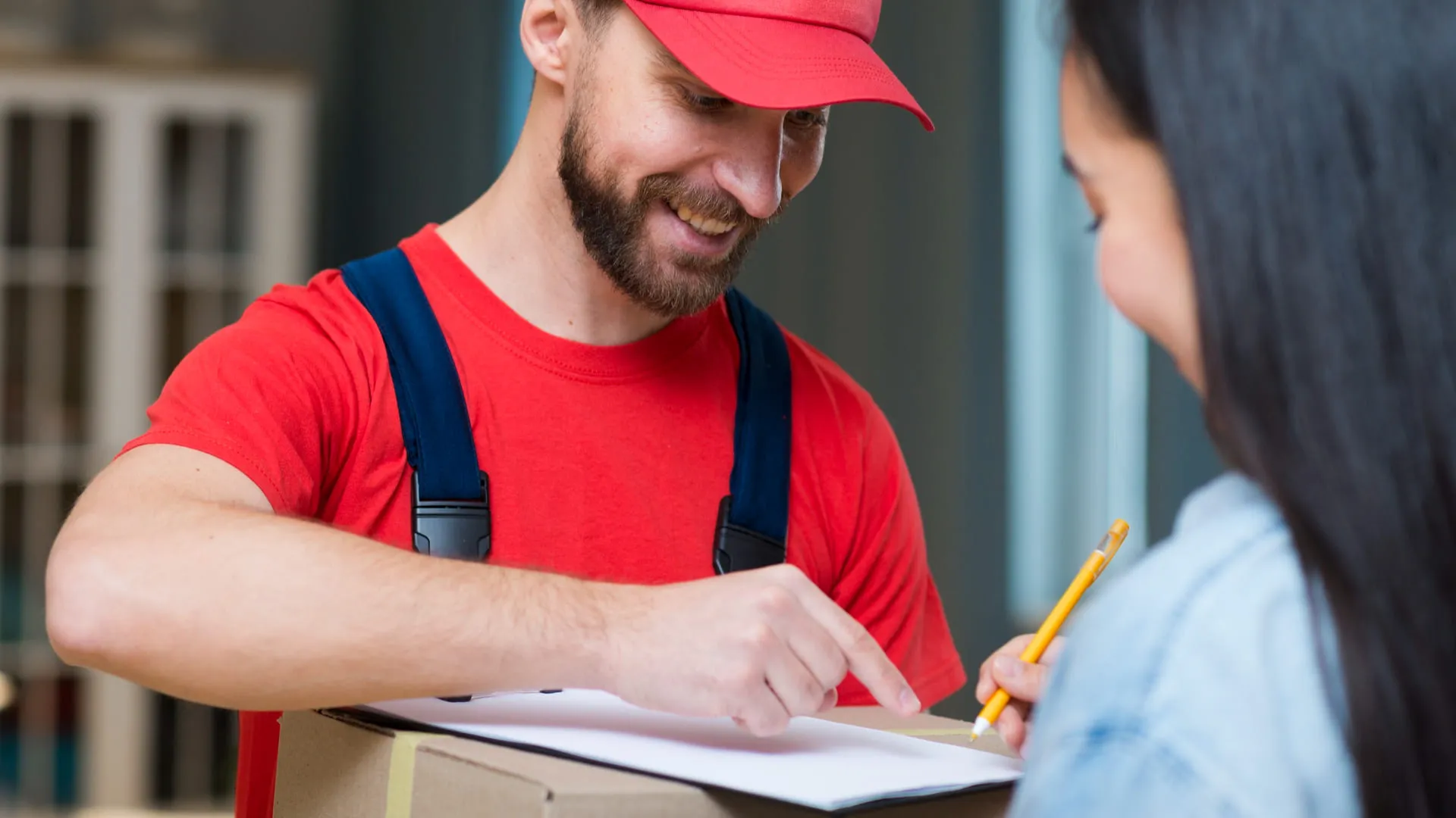 Man in red cap and shirt holding a box while woman signs paper on clipboard.