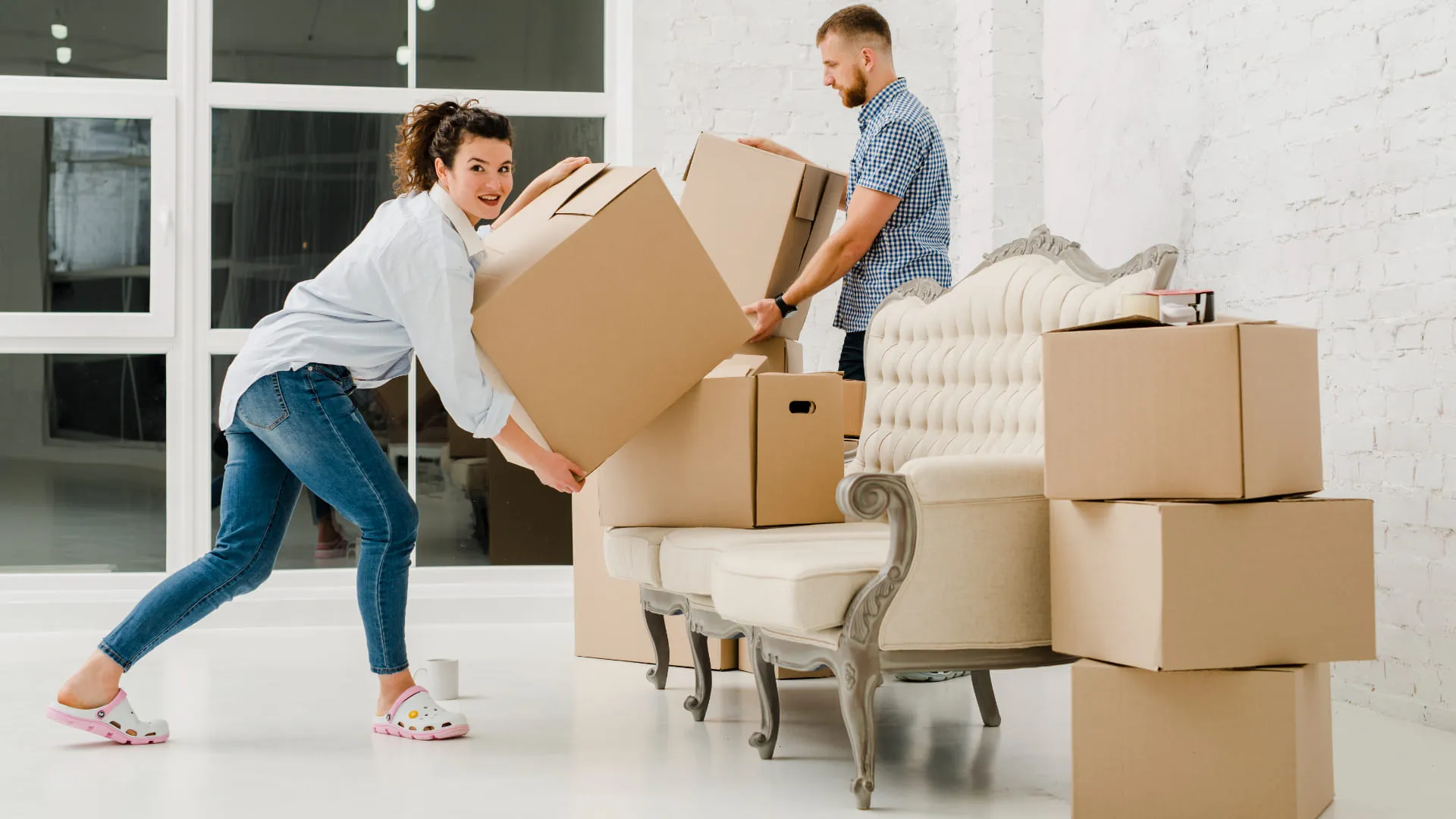 A woman and man carrying cardboard boxes in a bright room with packed furniture and boxes.