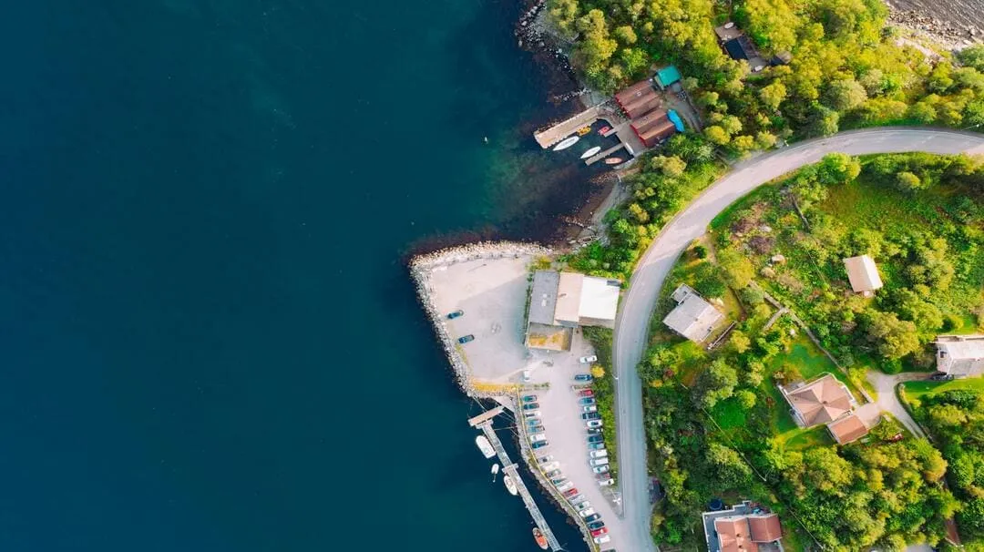 Aerial view of a curved road, green trees, houses, parking lot, docks, and deep blue water.