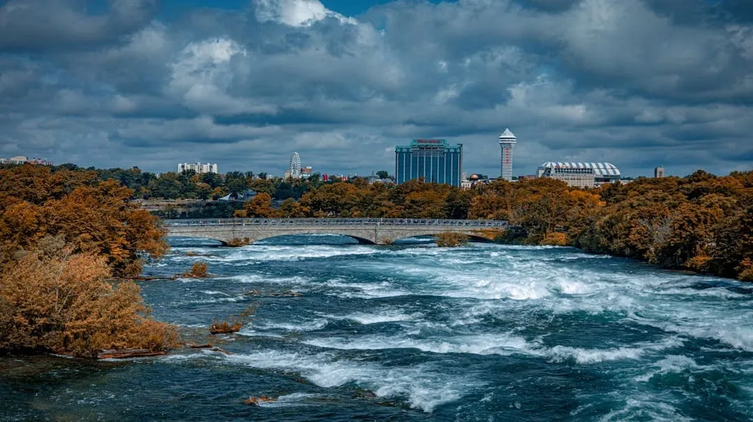 Rushing river with a stone bridge and autumn trees under a cloudy sky, with city buildings in the background.