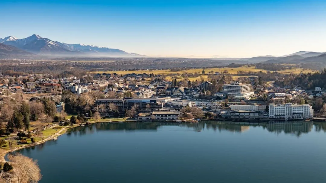 A lakeside town with buildings, trees, and mountains under a clear blue sky.