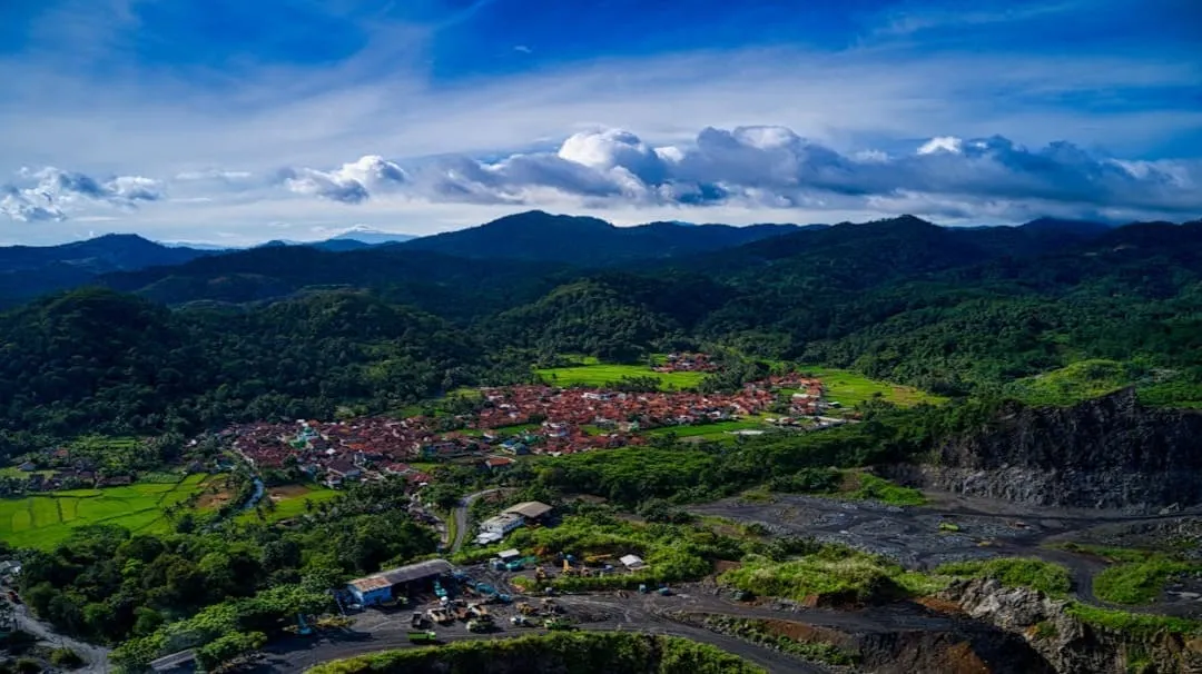 Aerial view of a village nestled among green hills under a blue sky with clouds.