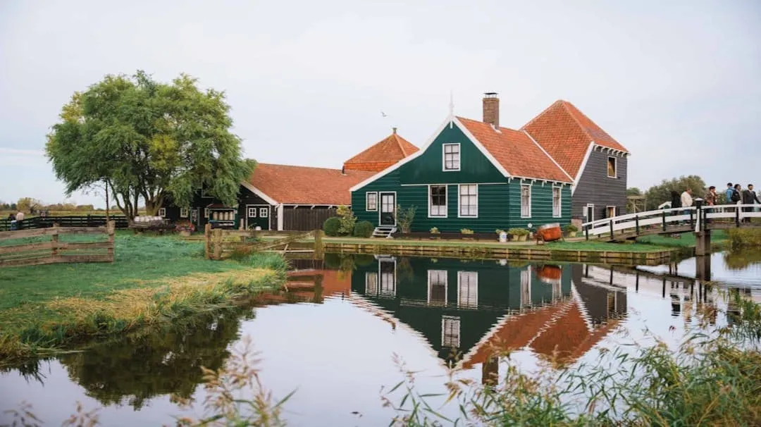 Green house with red roof beside calm water reflecting the building and a small white bridge nearby.