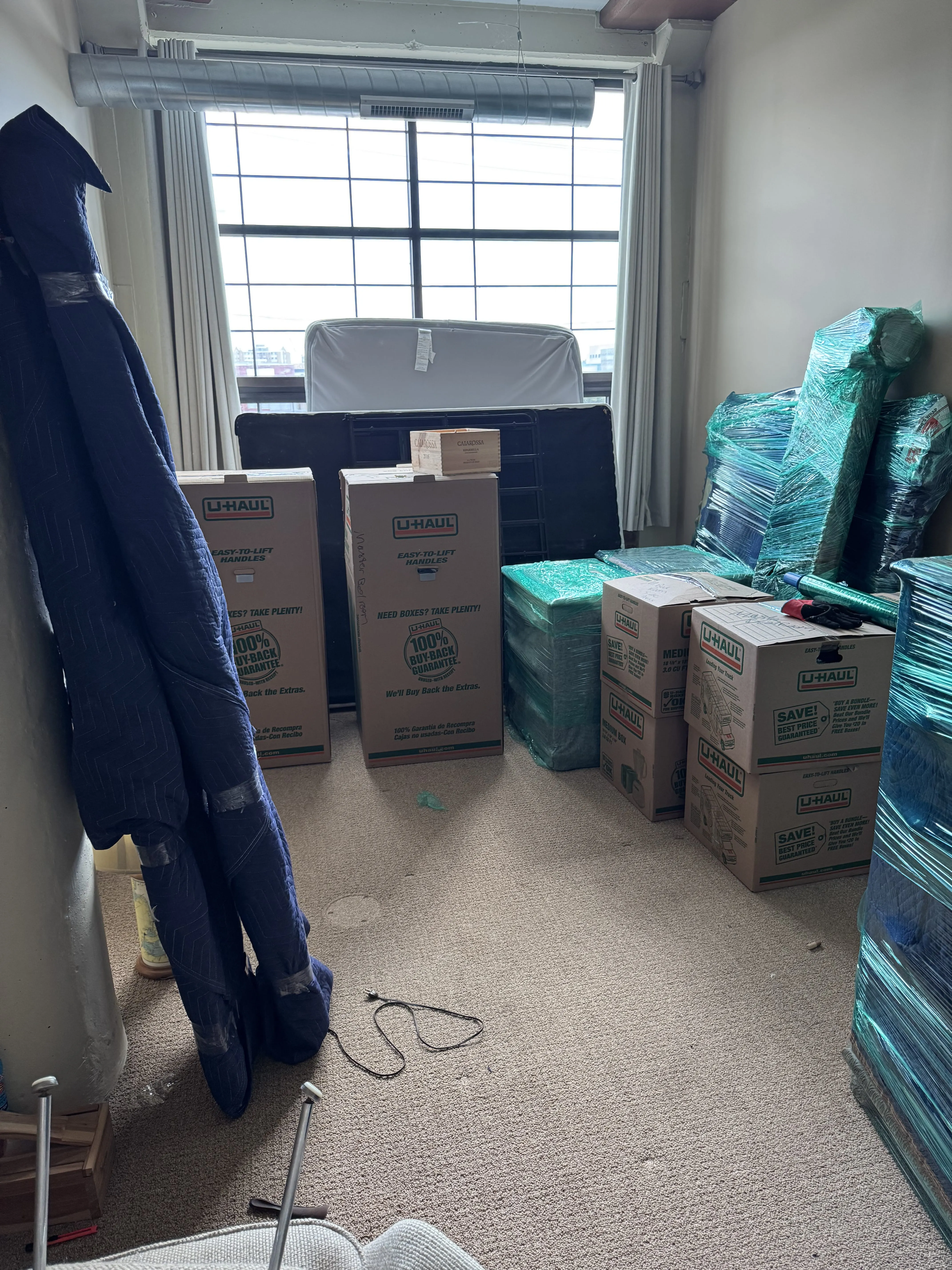 Room filled with U-Haul boxes, wrapped furniture, and moving blankets near a large window for local moving in Buffalo, NY