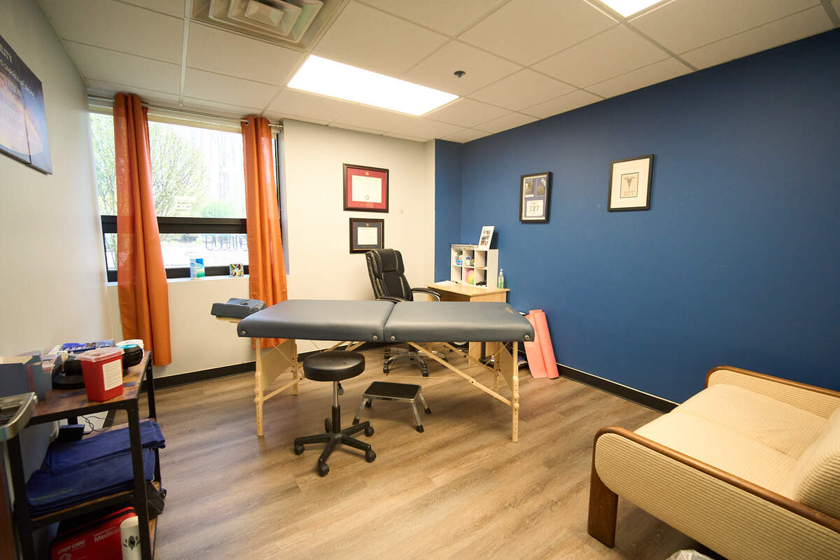 Bright massage room with treatment table, office chair, beige couch, wood floor, and blue accent wall.
