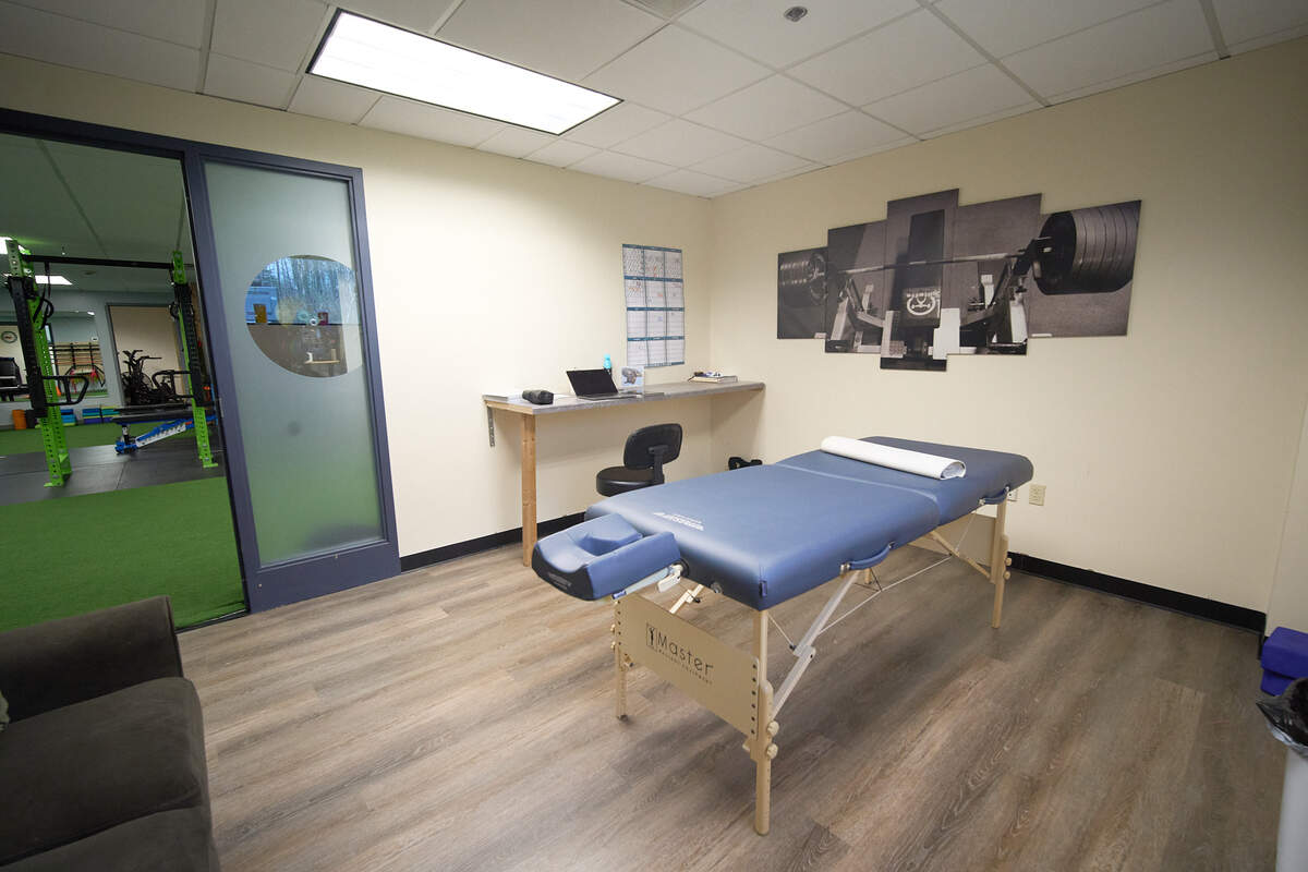Massage room with a blue massage table, desk with chair, and workout equipment visible through glass door.