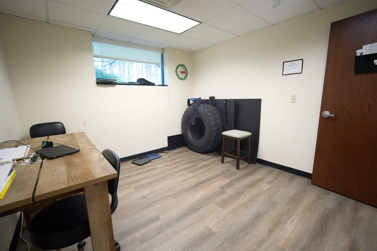 Small room with wooden desk, two black chairs, a window, large tire, stool, and wood door.
