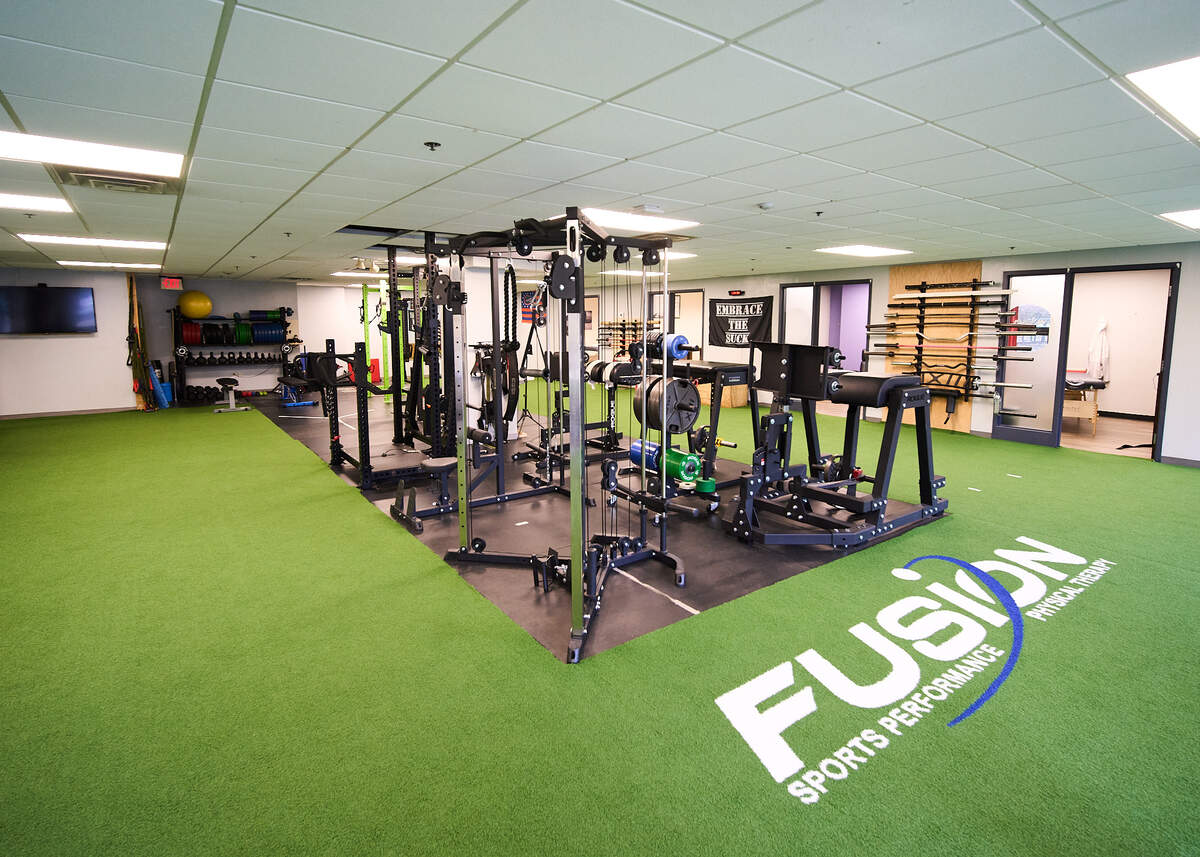 Sports performance gym with weight machines on black flooring and green turf with Fusion logo.