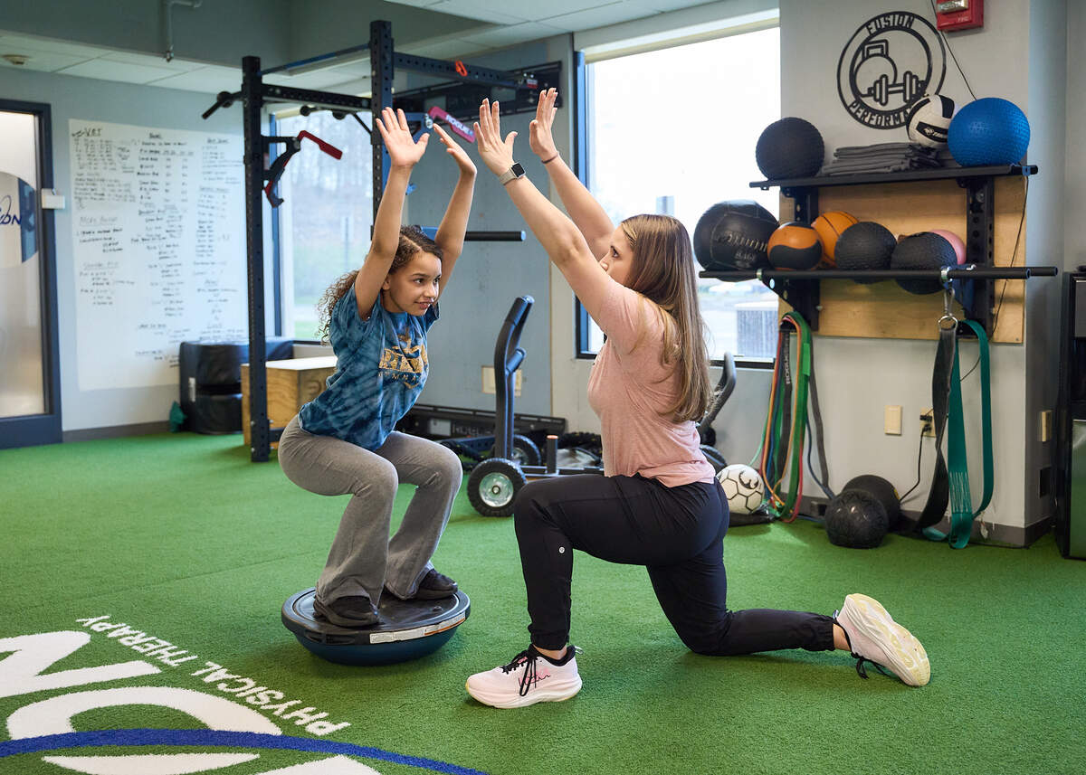 Young girl balancing on exercise ball while trainer kneels with arms raised facing her indoors.