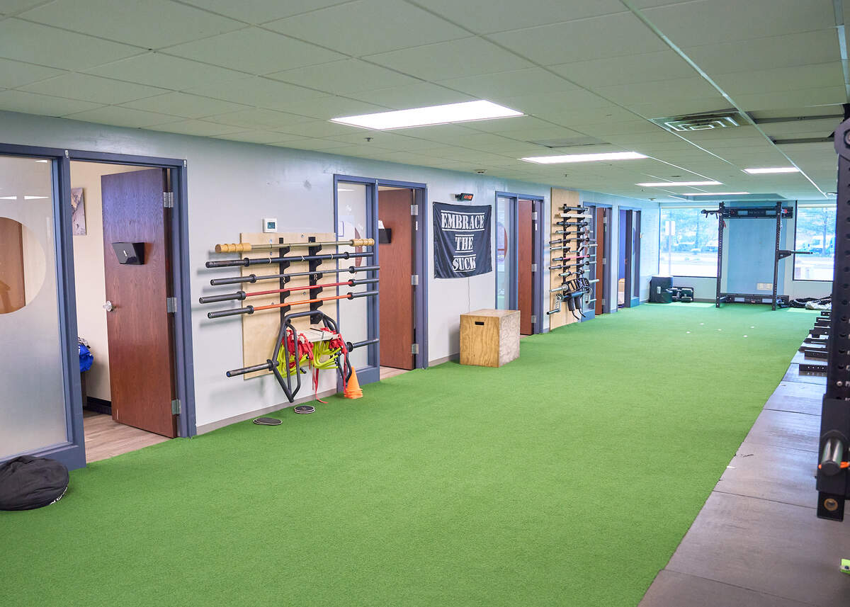 Indoor training room with green turf, barbells on racks, wooden plyo box, and large windows at the far end.