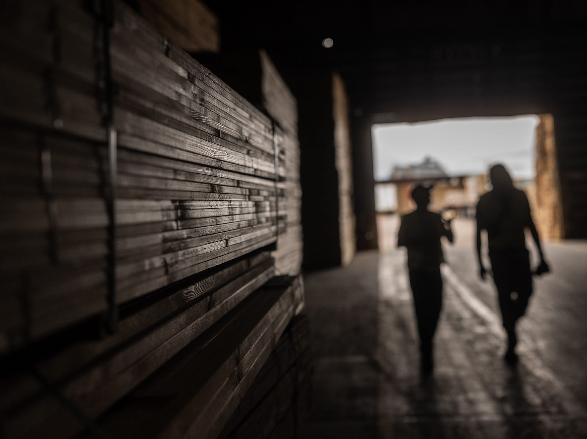 Two people silhoutted walking next to a stack of treated lumber.