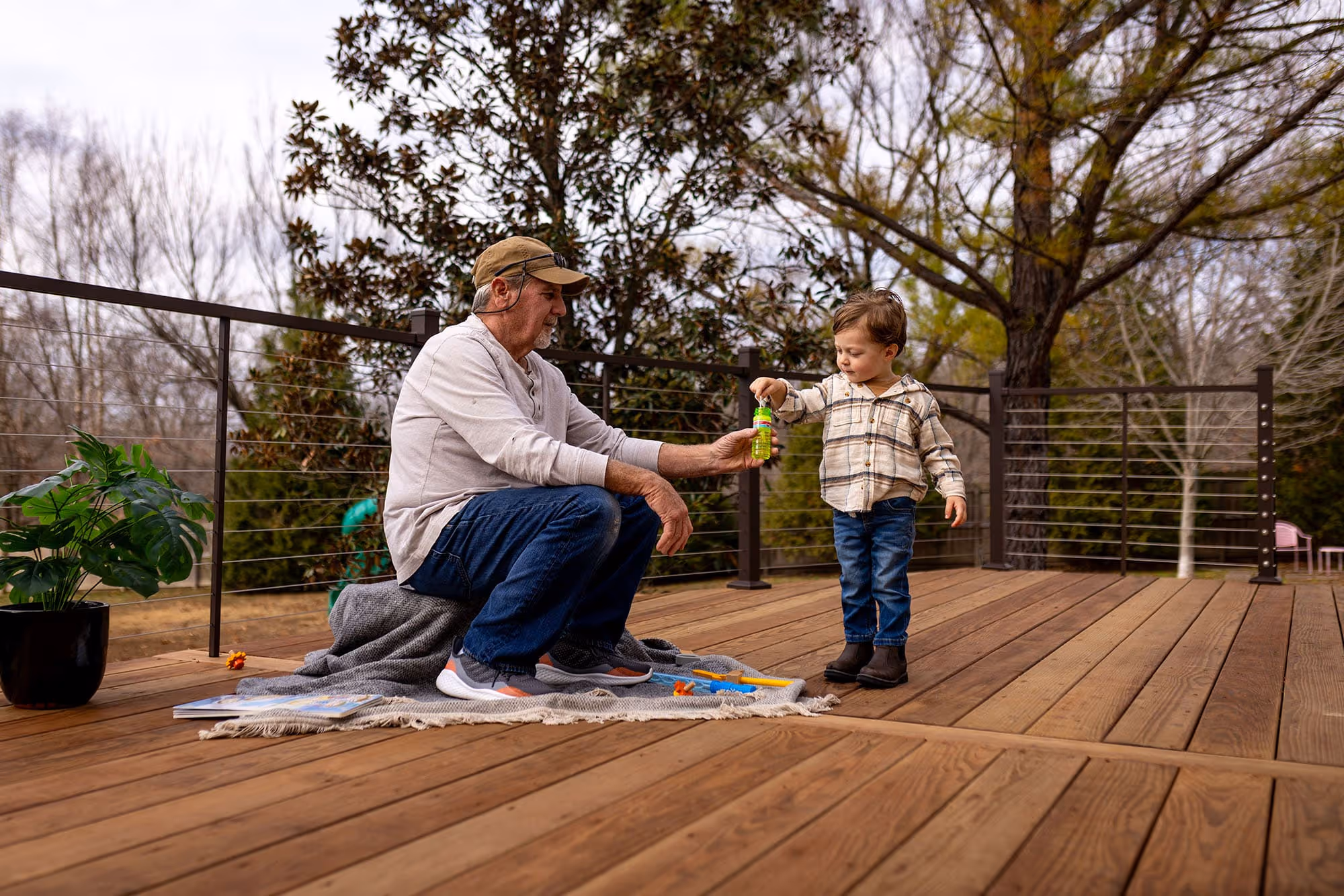 A person sitting down on a treated wood deck holding a container of bubbles as a kid dips the bubble stick in the container.