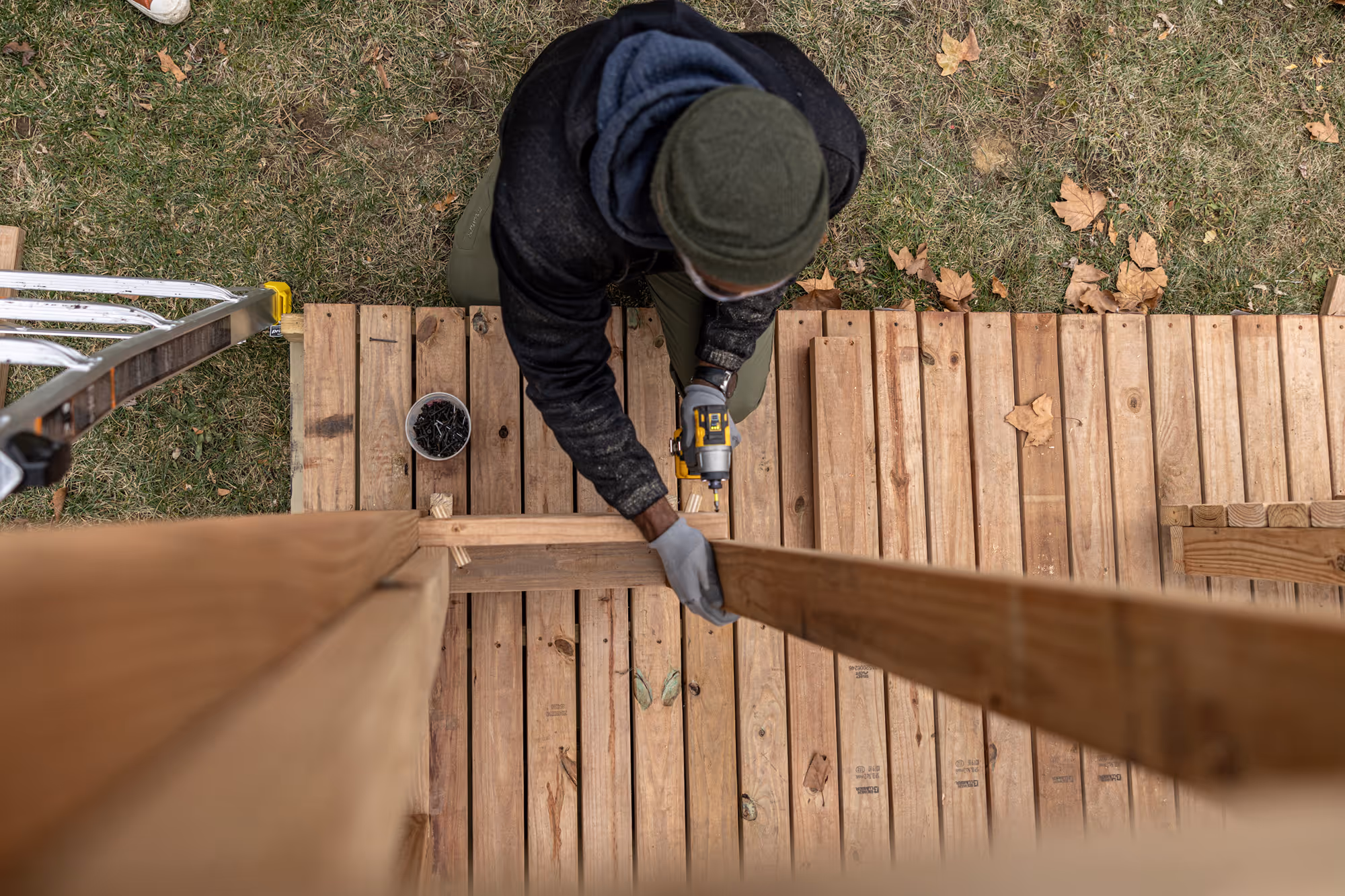 Person wearing a green beanie and gloves assembling a wooden deck using a power drill from an overhead perspective.