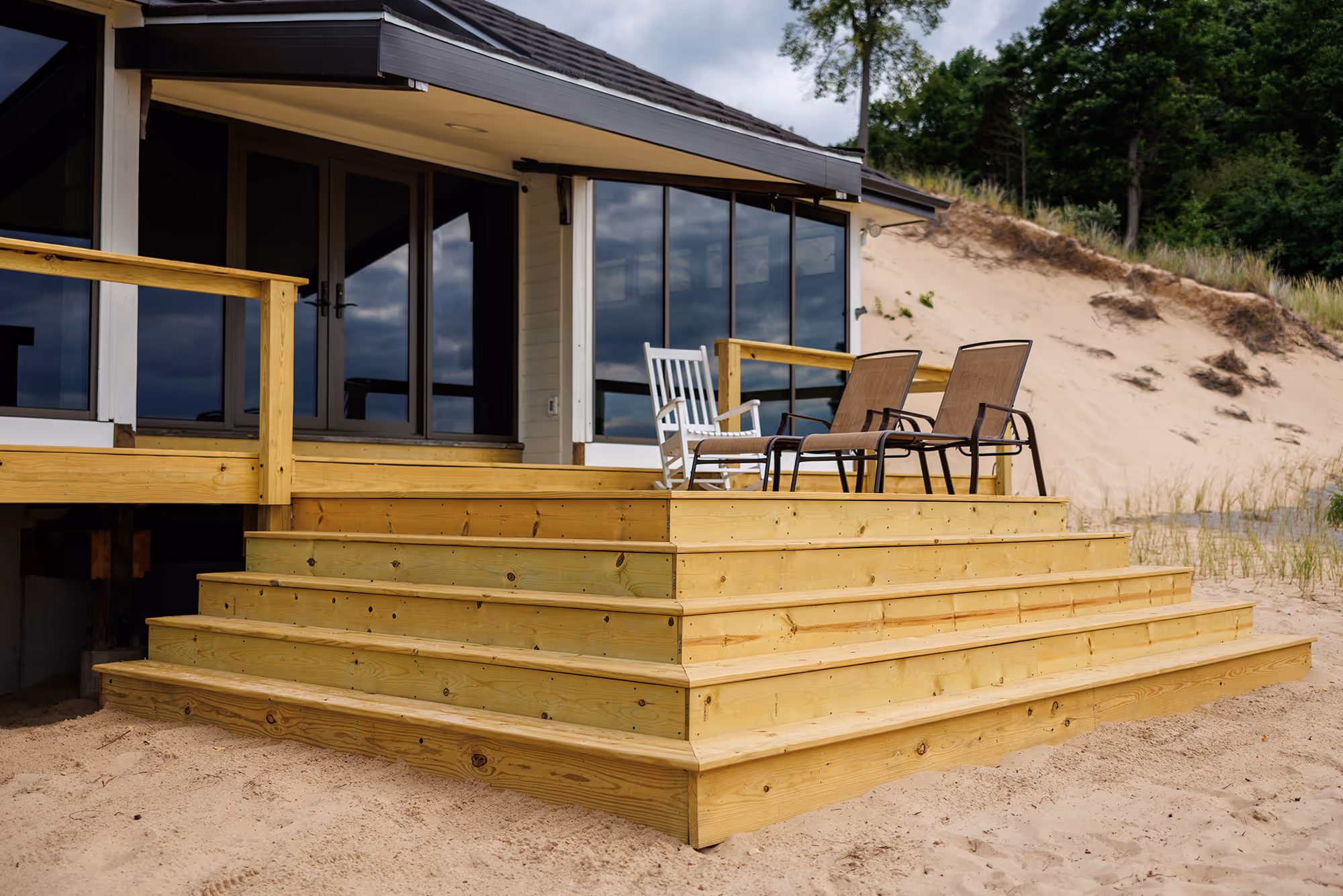 Pressure treated wooden deck stairs leading to a porch with two brown chairs and a white rocking chair, in front of a house near sandy dunes and trees.