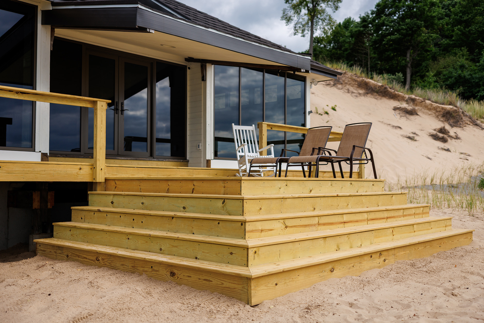 Pressure treated wooden deck stairs leading to a porch with two brown chairs and a white rocking chair, in front of a house near sandy dunes and trees.