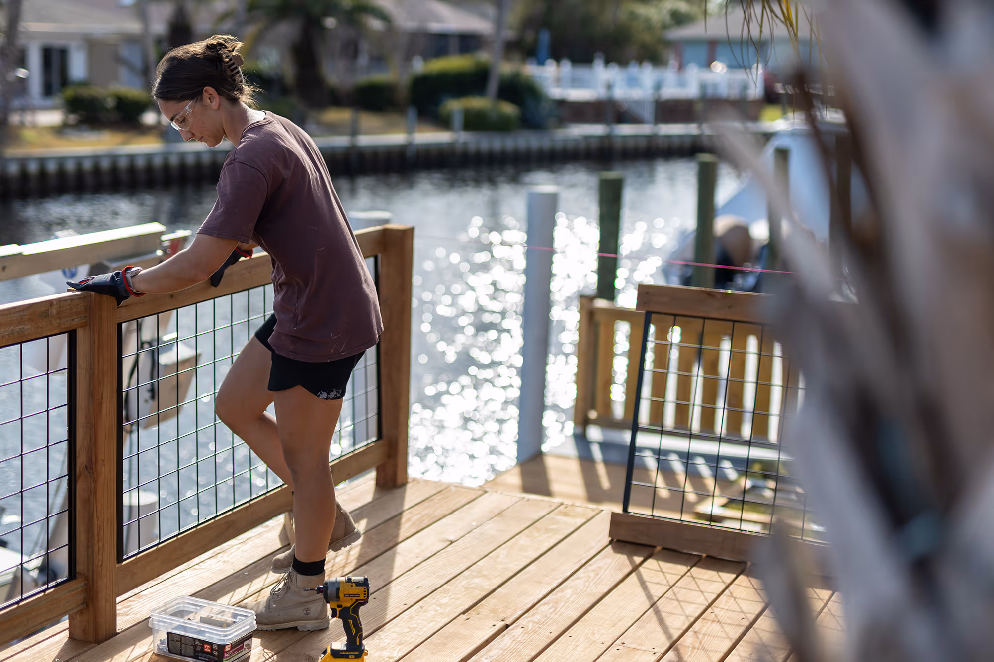 Person wearing safety glasses and gloves installing a wooden railing on a deck overlooking water.