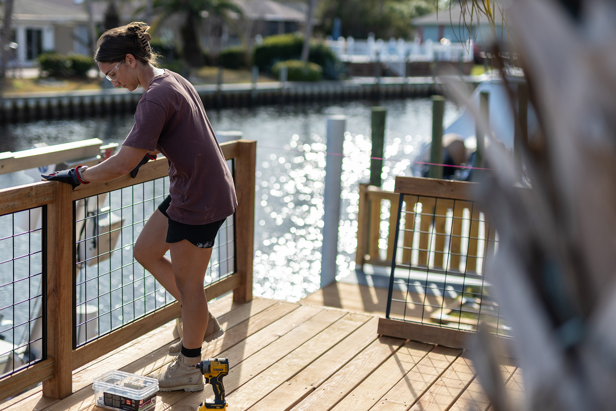 Person wearing safety glasses and gloves installing a wooden railing on a deck overlooking water.