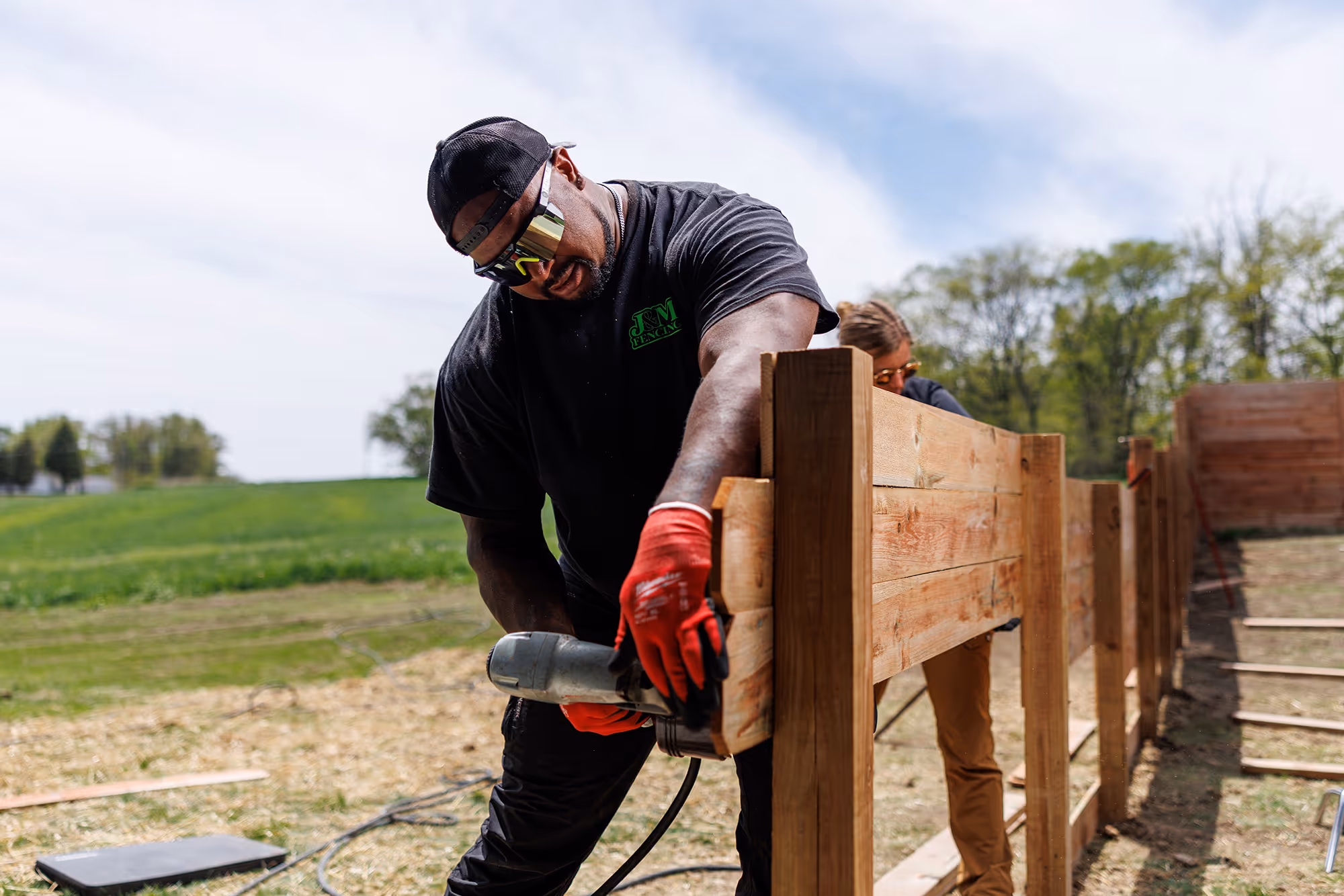 Construction worker wearing red gloves and safety glasses cutting wooden fence boards outdoors.