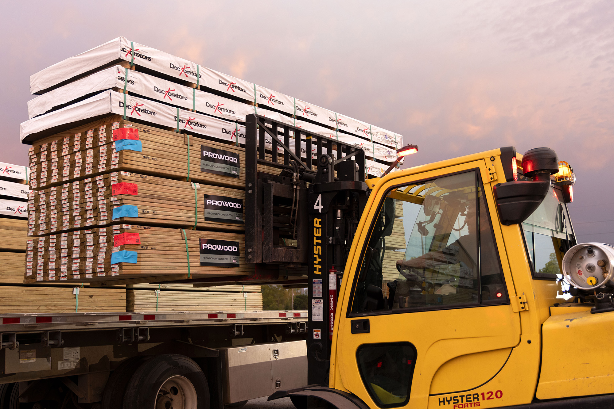 Yellow forklift loading stacked ProWood lumber and Deckorators decking wrapped in white protective covers onto a flatbed truck at sunset.